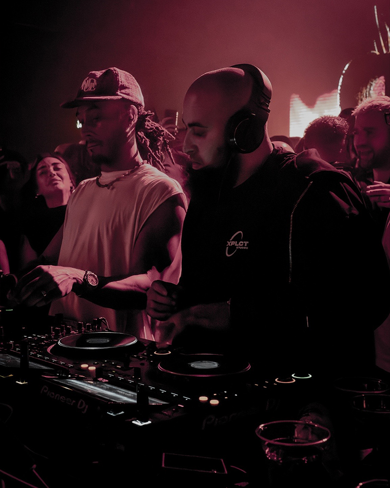 AfroTura behind the Pioneer DJ decks during a live club performance, surrounded by an energetic crowd under red and pink lighting