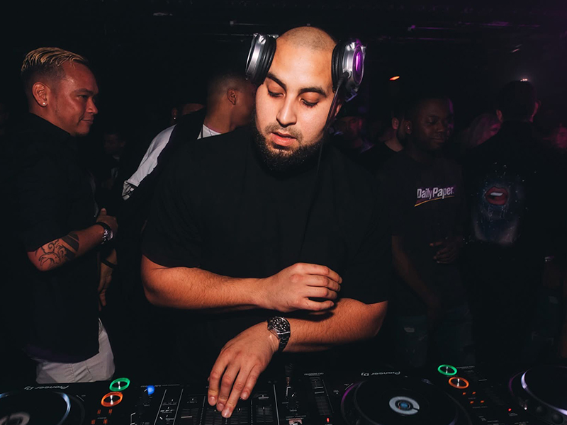 AfroTura concentrated behind Pioneer DJ decks during a packed club night, wearing headphones and a black t-shirt with an energetic crowd behind him