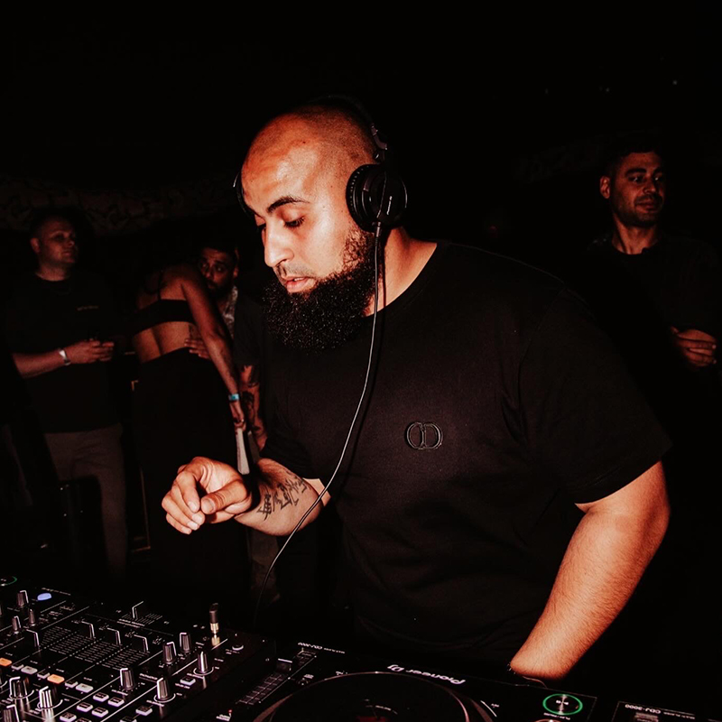 AfroTura focused behind Pioneer DJ decks during a late-night club performance, wearing headphones and a black Dior t-shirt with crowd in the background.