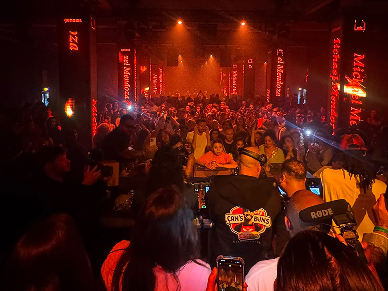 Wide shot of AfroTura behind the decks at a large club event with LED displays reading Michael Mendoza, facing a packed crowd under red atmospheric lighting