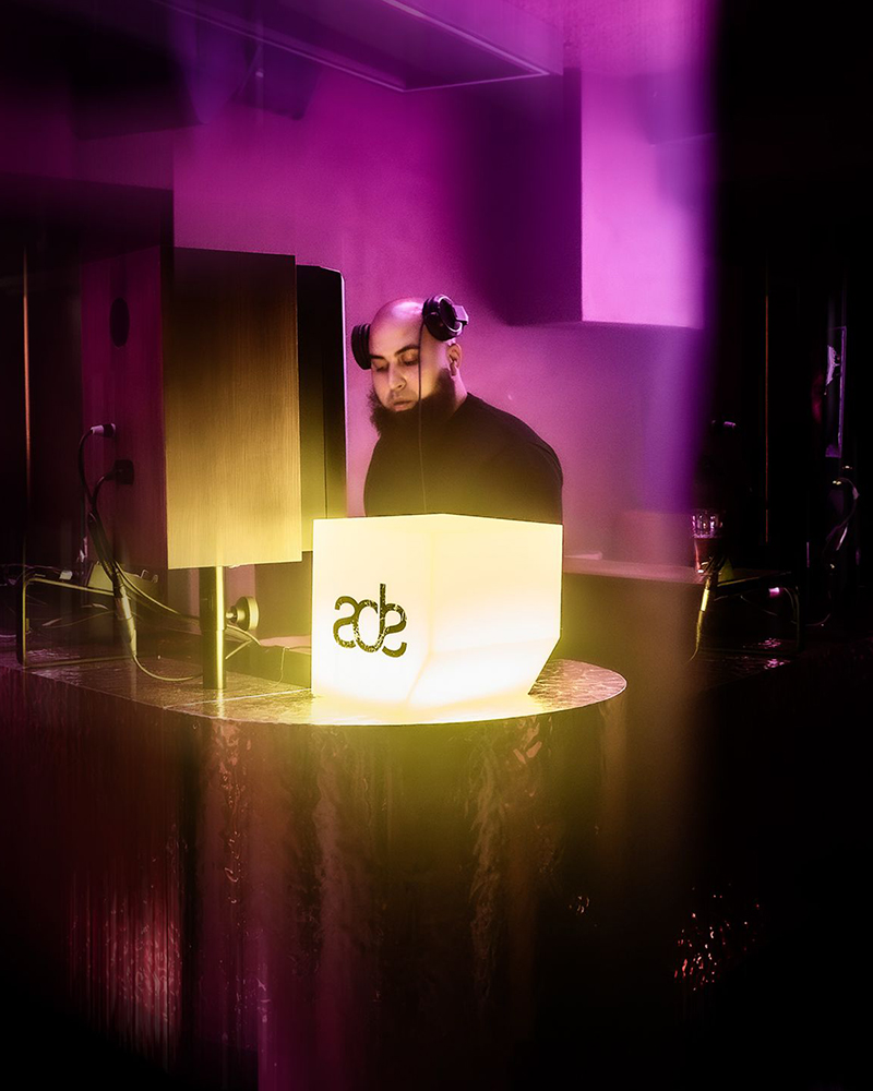AfroTura performing at Amsterdam Dance Event (ADE), focused behind the decks with a glowing ADE cube and purple atmospheric lighting