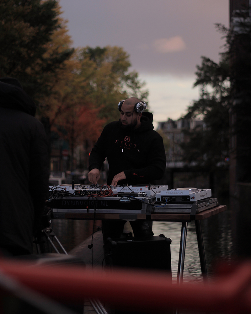 AfroTura performing a DJ set on the banks of an Amsterdam canal at dusk, surrounded by autumn foliage