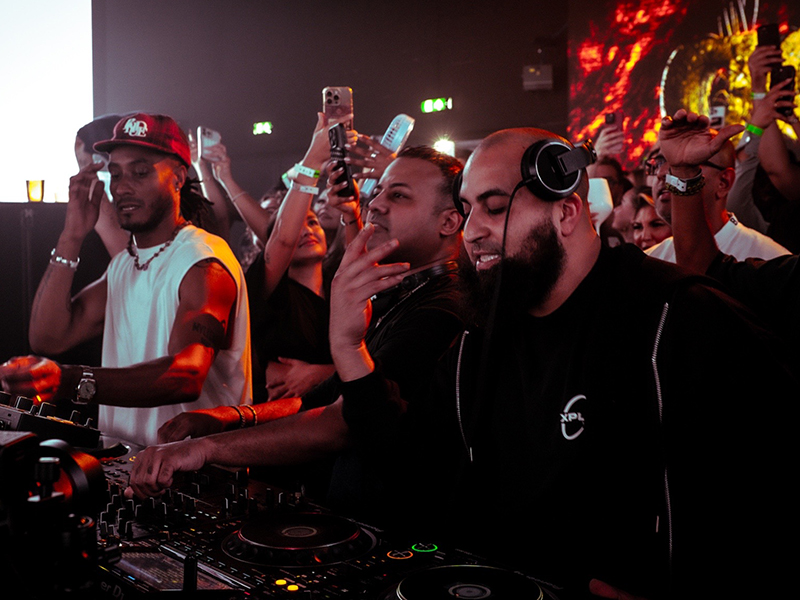 AfroTura performing an intense DJ set in a packed club under warm orange lighting, with an excited crowd holding up phones and filming