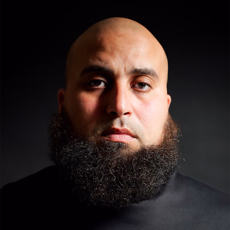 Close-up portrait of a bald man with a thick, curly black beard and a serious expression against a dark background.