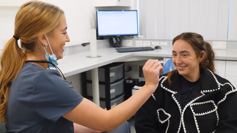 Female dental professional smiling and showing a clear dental aligner to a seated woman in a clinic.