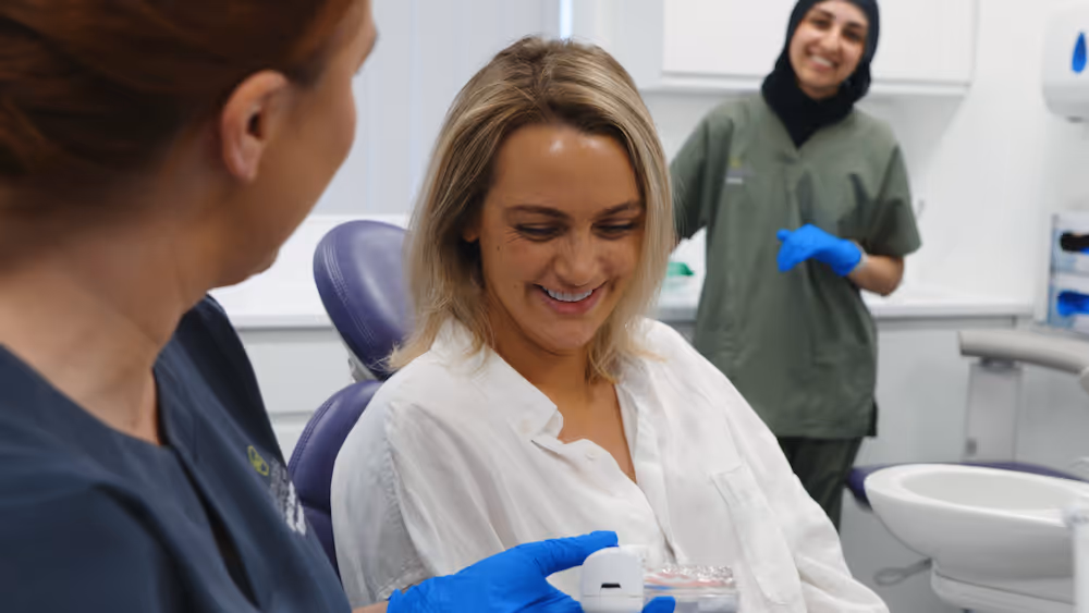A dental patient smiling while a dental professional shows her a dental model, with another staff member watching in the background in a clinic.