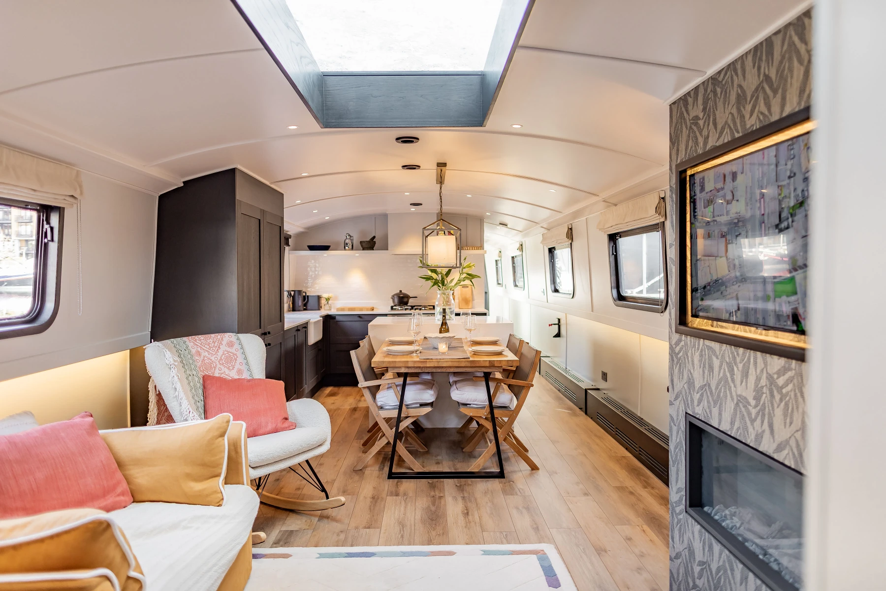 Interior of a modern narrowboat with a dining table set for four, a kitchen with dark cabinets, and a cozy seating area with colorful cushions.
