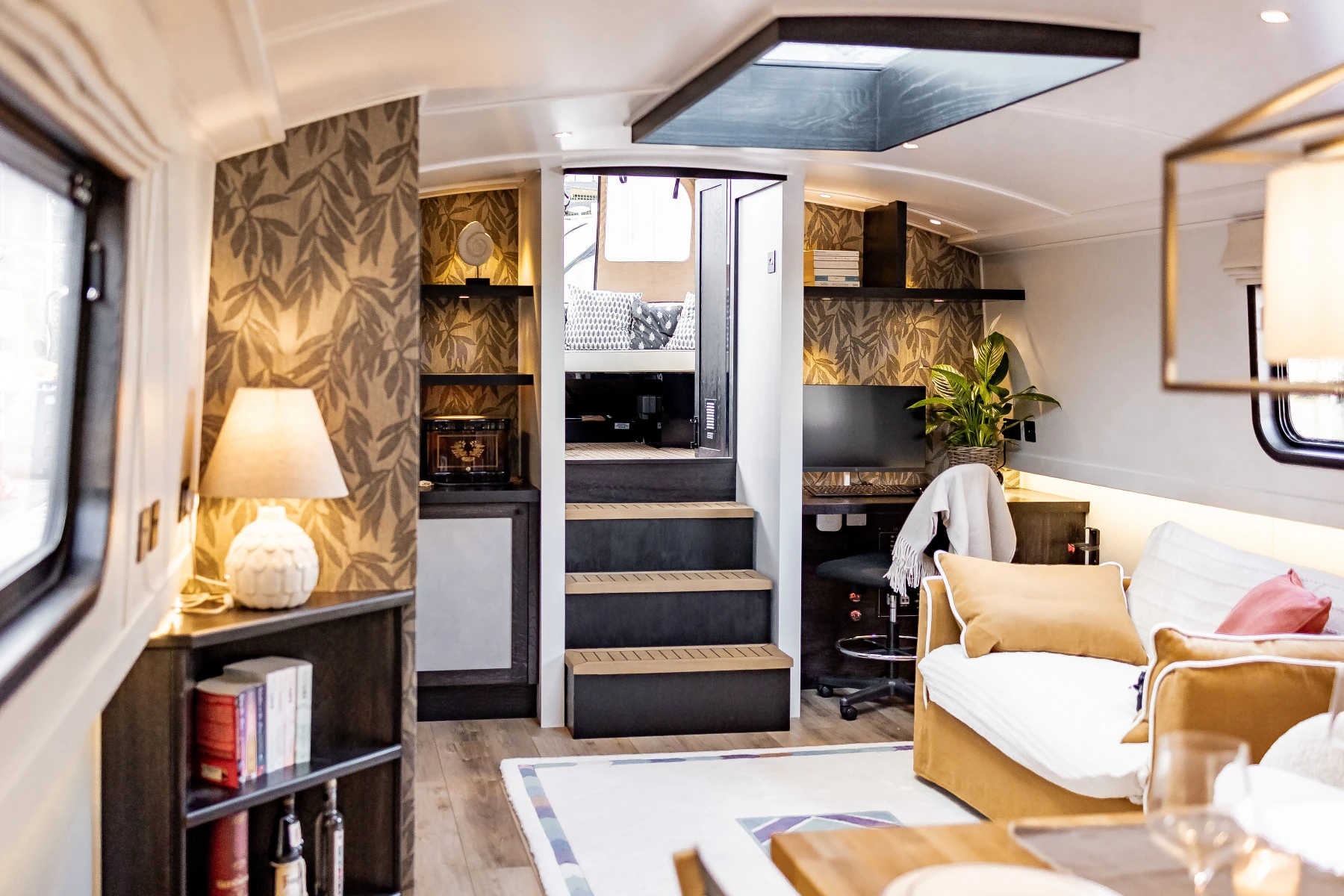 Cozy living space with mustard sofa, decorative pillows, desk with chair, potted plant, patterned wallpaper, lamp, and stairs leading to a small seating area.