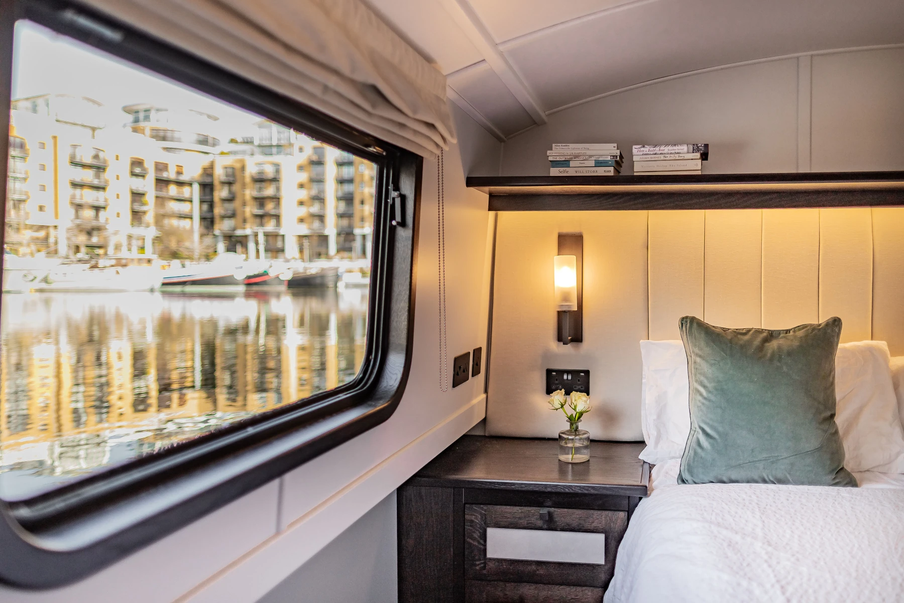 Cozy bedroom corner with a green pillow, white bedding, wooden nightstand with flowers, lit wall lamp, and a window showing waterfront buildings and boats.