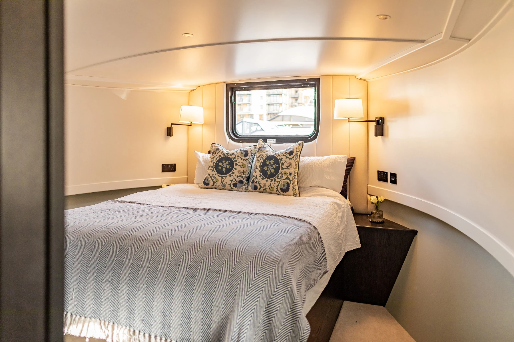 Cozy bedroom with a large bed, decorative pillows, side lamps, and a window overlooking buildings.