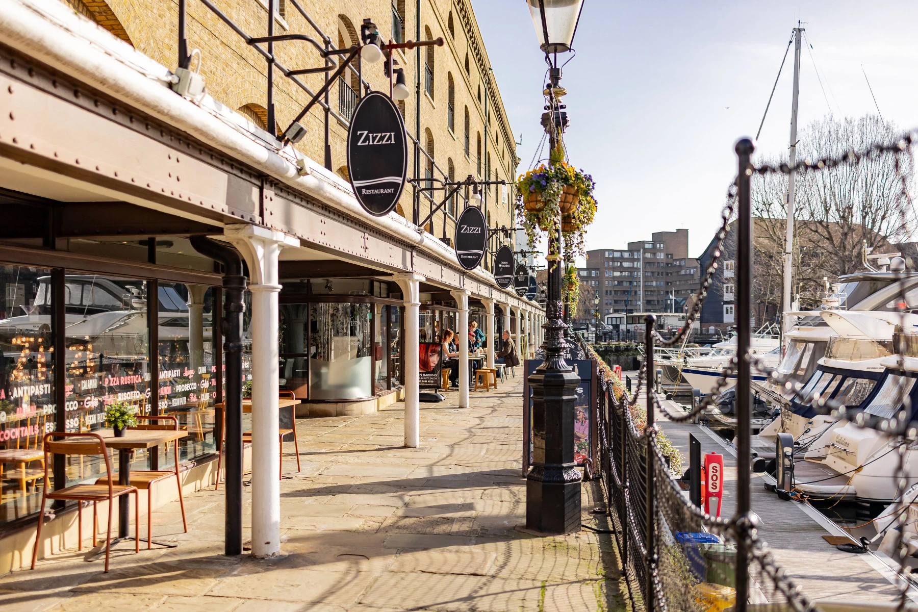 Outdoor seating area of Zizzi restaurant along a waterfront with boats docked nearby under a clear sky.