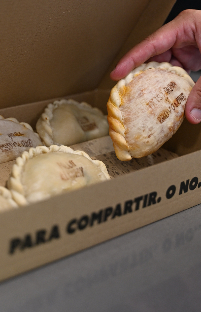 Hand holding an empanada with inscription inside a cardboard box with more empanadas.