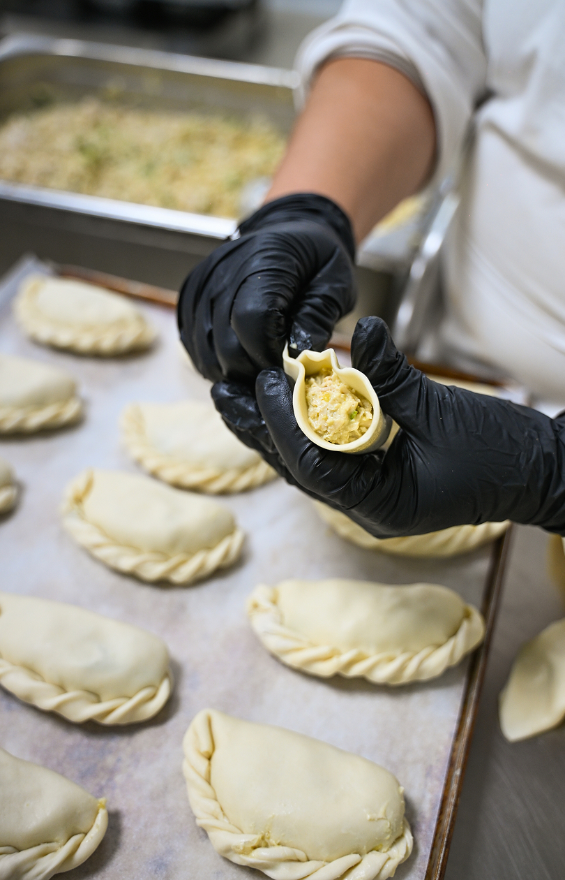 Hands with black gloves folding dough around filling to make empanadas on a baking tray.
