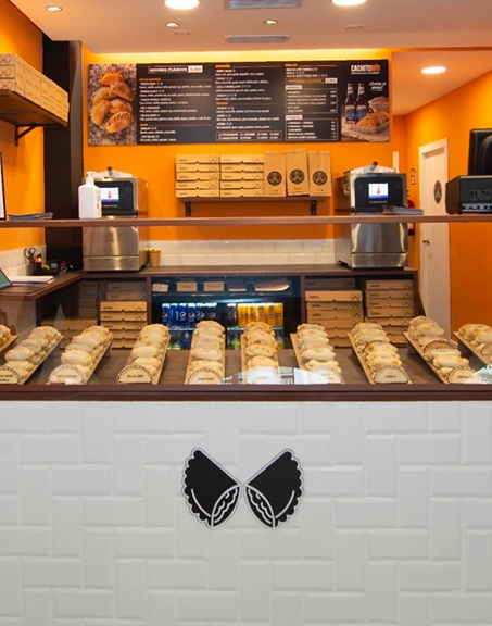 Display of assorted empanadas in trays at a bright orange bakery counter with menu boards above.