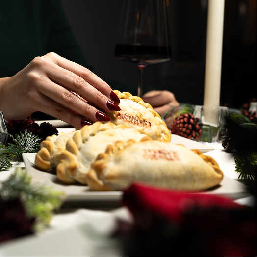 Hand with red nails reaching for a cheeseburger empanada on a festive table with wine and pine cones.