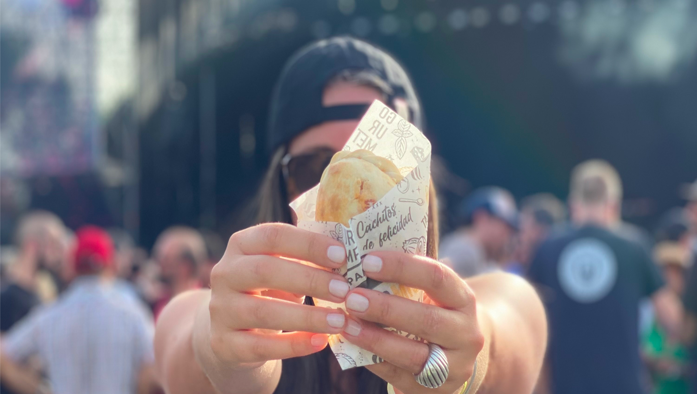 Person wearing sunglasses and a cap holding a wrapped empanada towards the camera at an outdoor event.