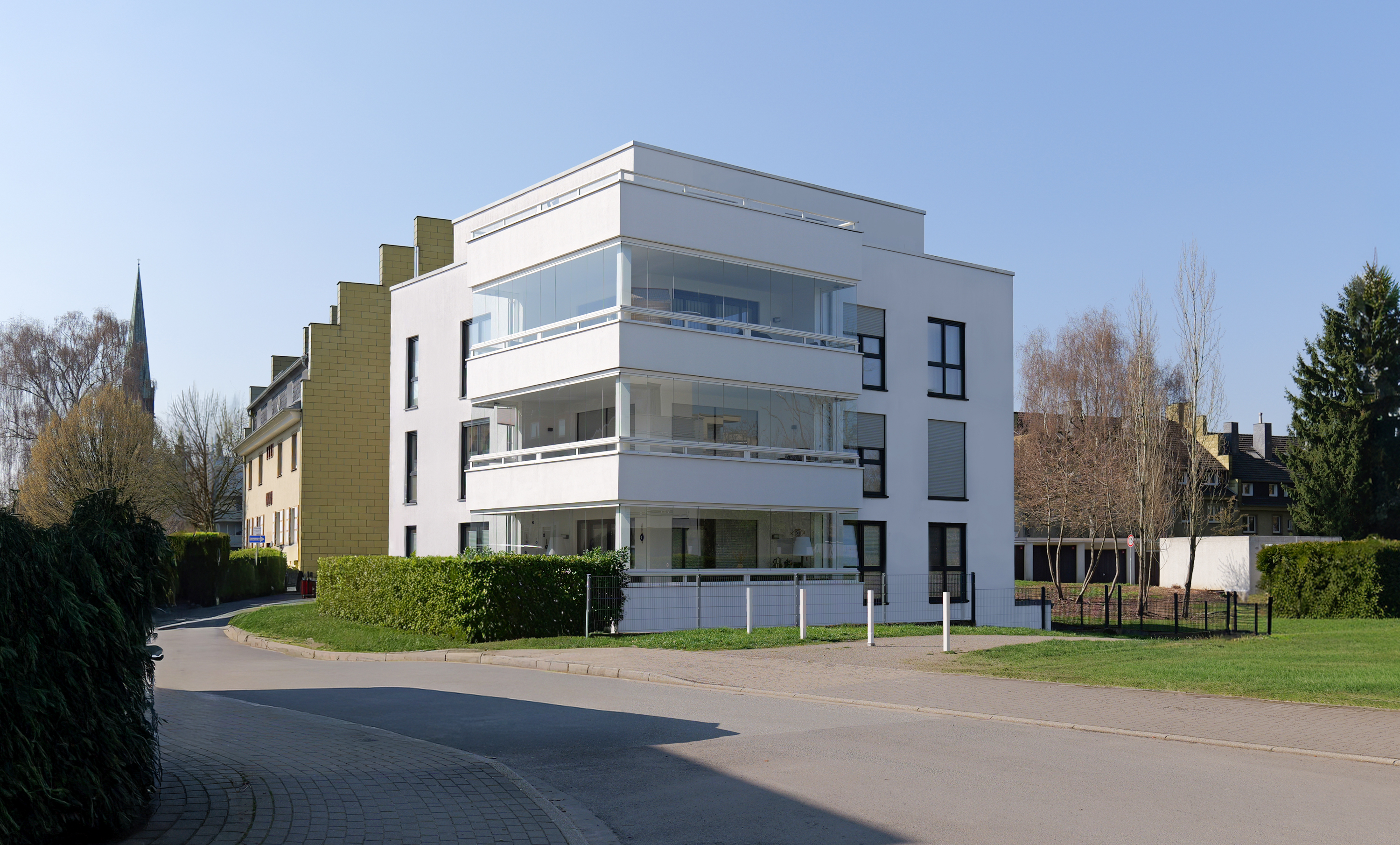 Modern weißes Wohngebäude mit großzügig verglasten Loggien in ruhiger Straße bei blauem Himmel.