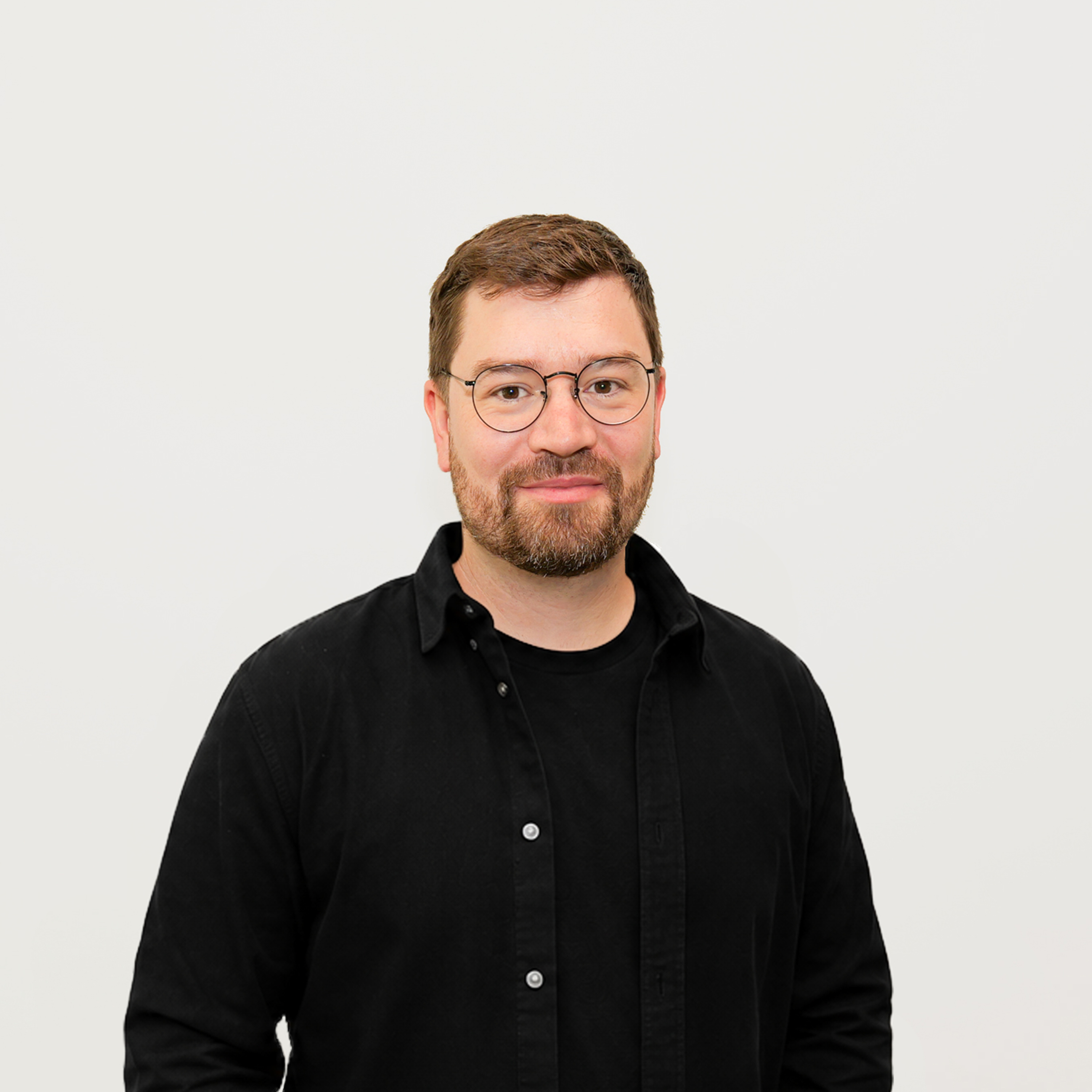 Mann mit braunen Haaren, Brille und Bart in schwarzem Hemd vor weißem Hintergrund.