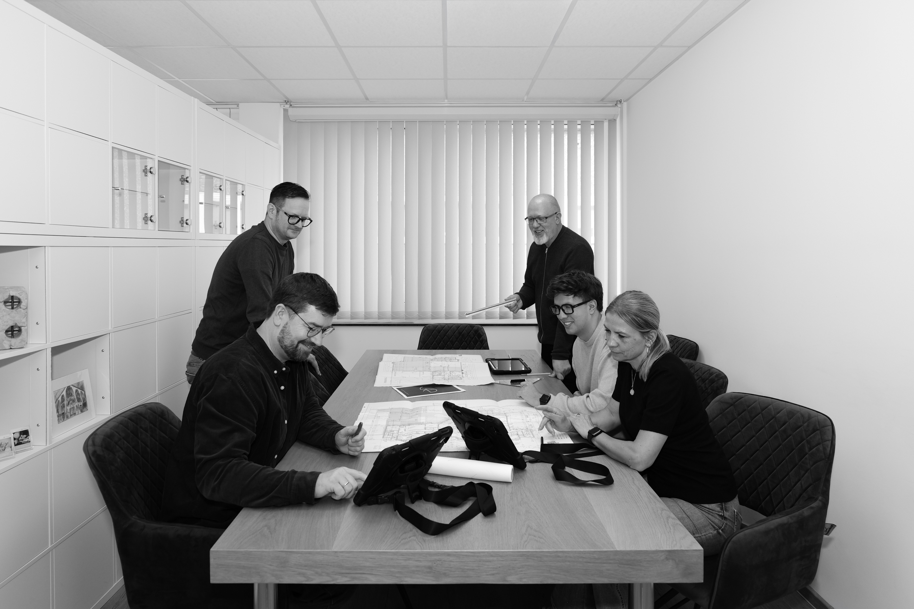 Fünf Personen besprechen Pläne an einem Tisch in einem modernen Büro mit Tablet und Zeichnungen.