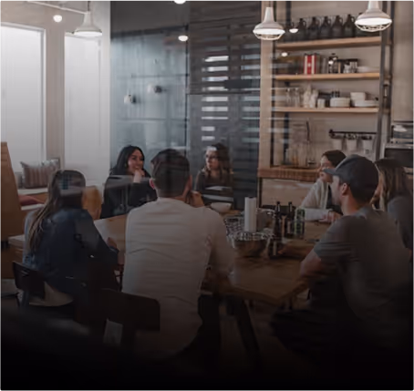 Six people sitting around a wooden table in a modern meeting room with shelves and hanging lights.