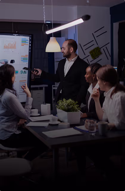 Man in suit giving presentation to three seated colleagues in a dimly lit modern office.