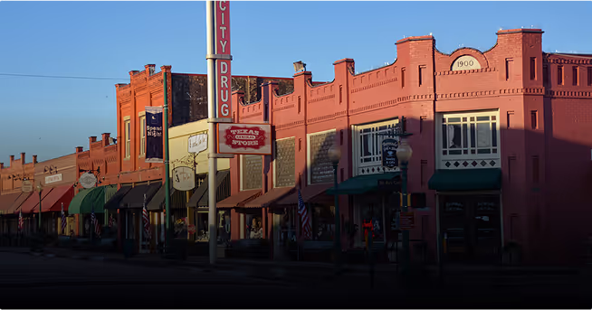 Historic red brick buildings with signs including City Drug and Texas Cross Store under clear sky.