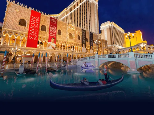 Venetian hotel and casino at night with gondolas on blue water and a white arched bridge.