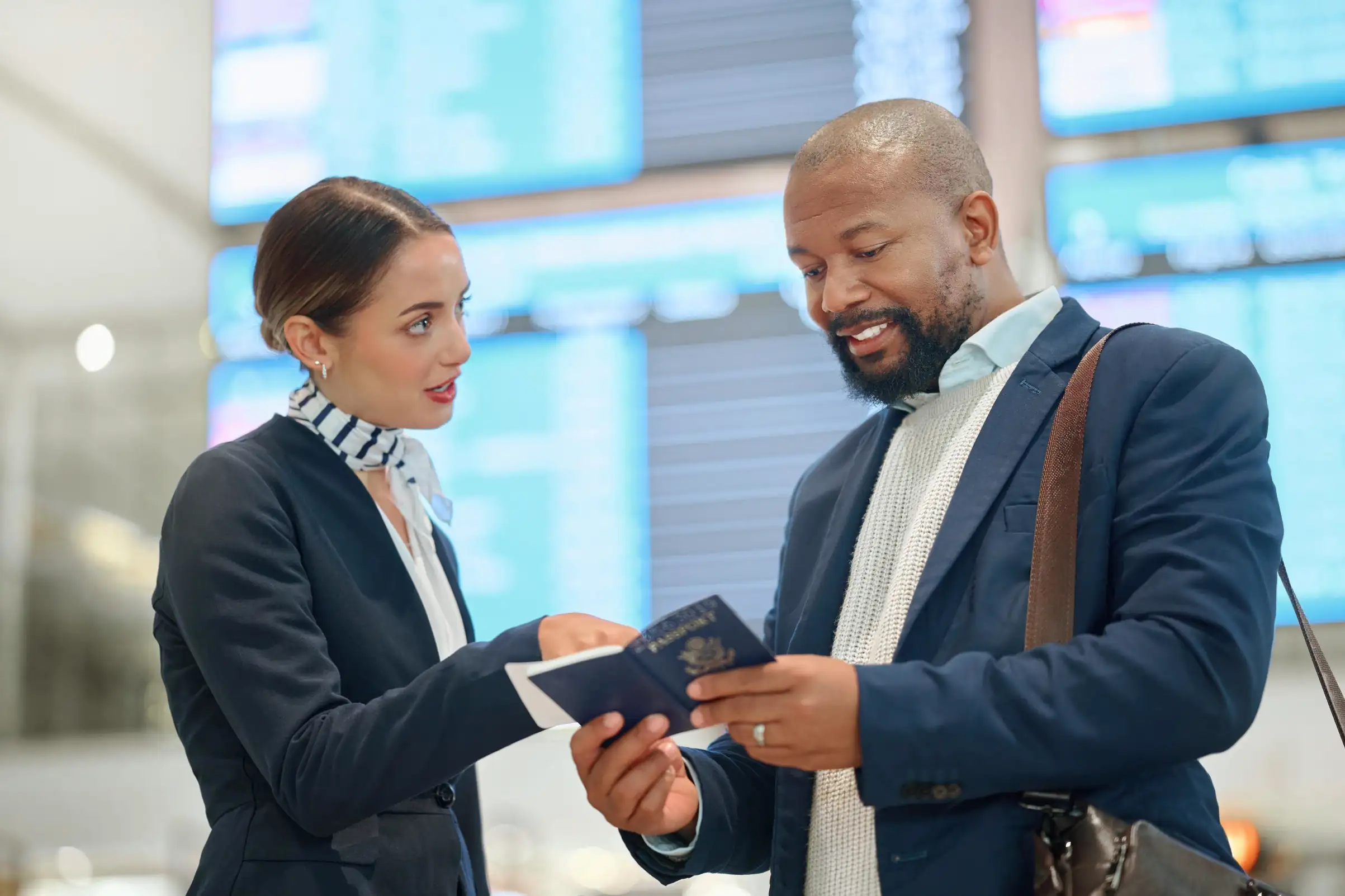 Airport staff assisting male traveler with passport and boarding pass near flight information screens.