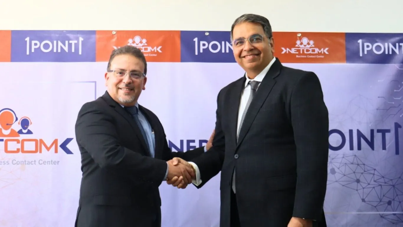 Two men in suits shaking hands in front of a business backdrop with logos and text.