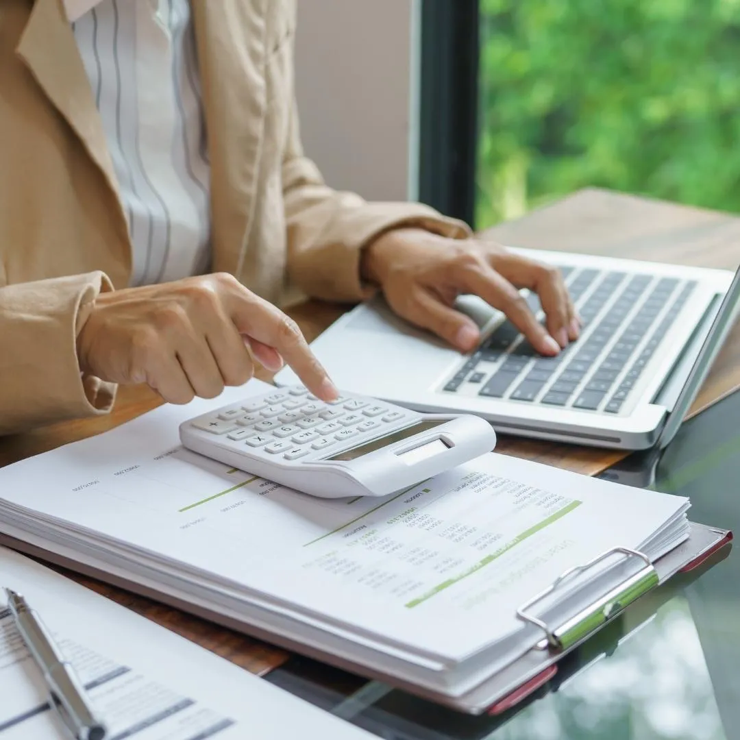 Person using a calculator and laptop to analyze financial documents on a clipboard.