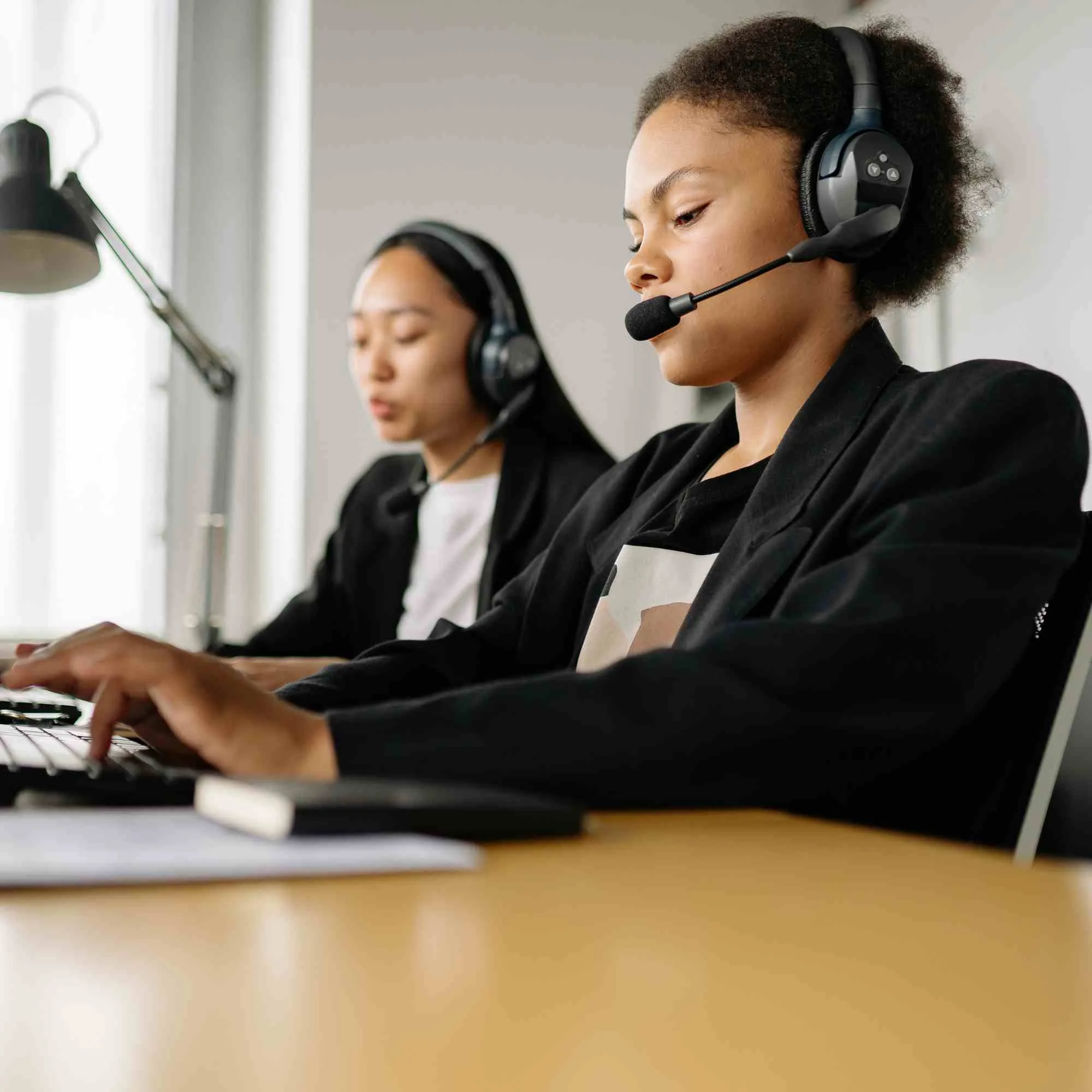 Two women wearing headsets working on computers in a bright office setting.