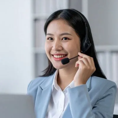Smiling woman with long dark hair wearing a headset and light blue blazer, speaking while holding the microphone near her mouth.