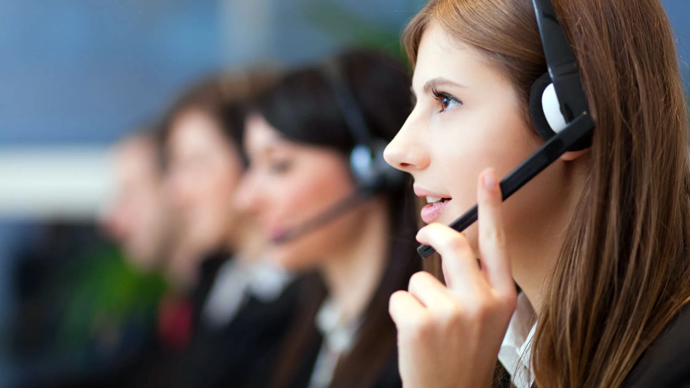 Close-up of a woman with brown hair wearing a headset and speaking, with three blurred colleagues also wearing headsets in the background, indicating a call center or customer support environment.