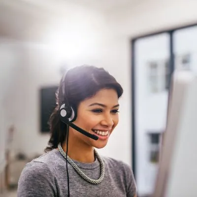 Smiling woman wearing a headset and gray sweater, working at a computer in a bright office.