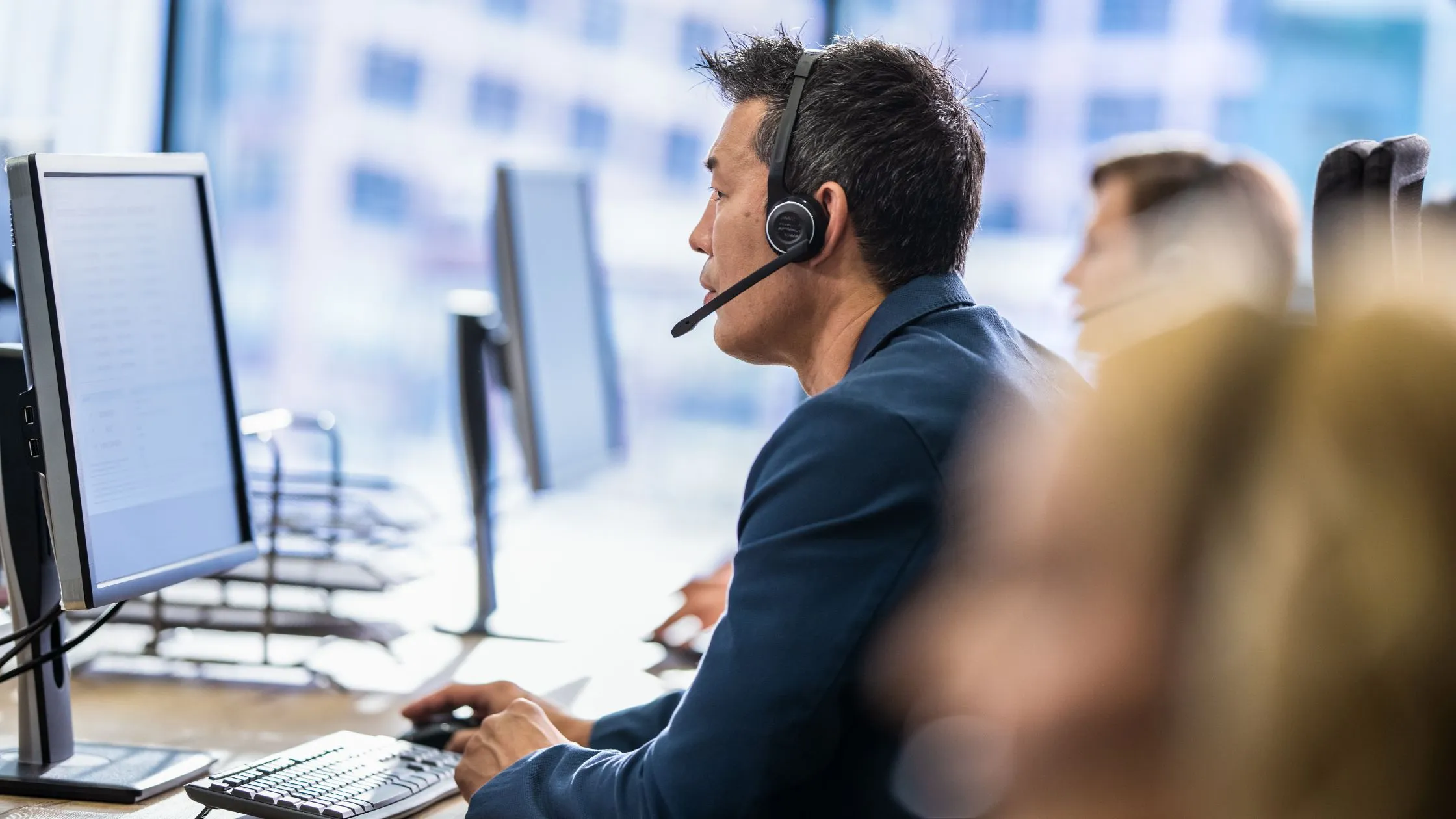 Man wearing a headset working at a computer in an office with other workers in the background.