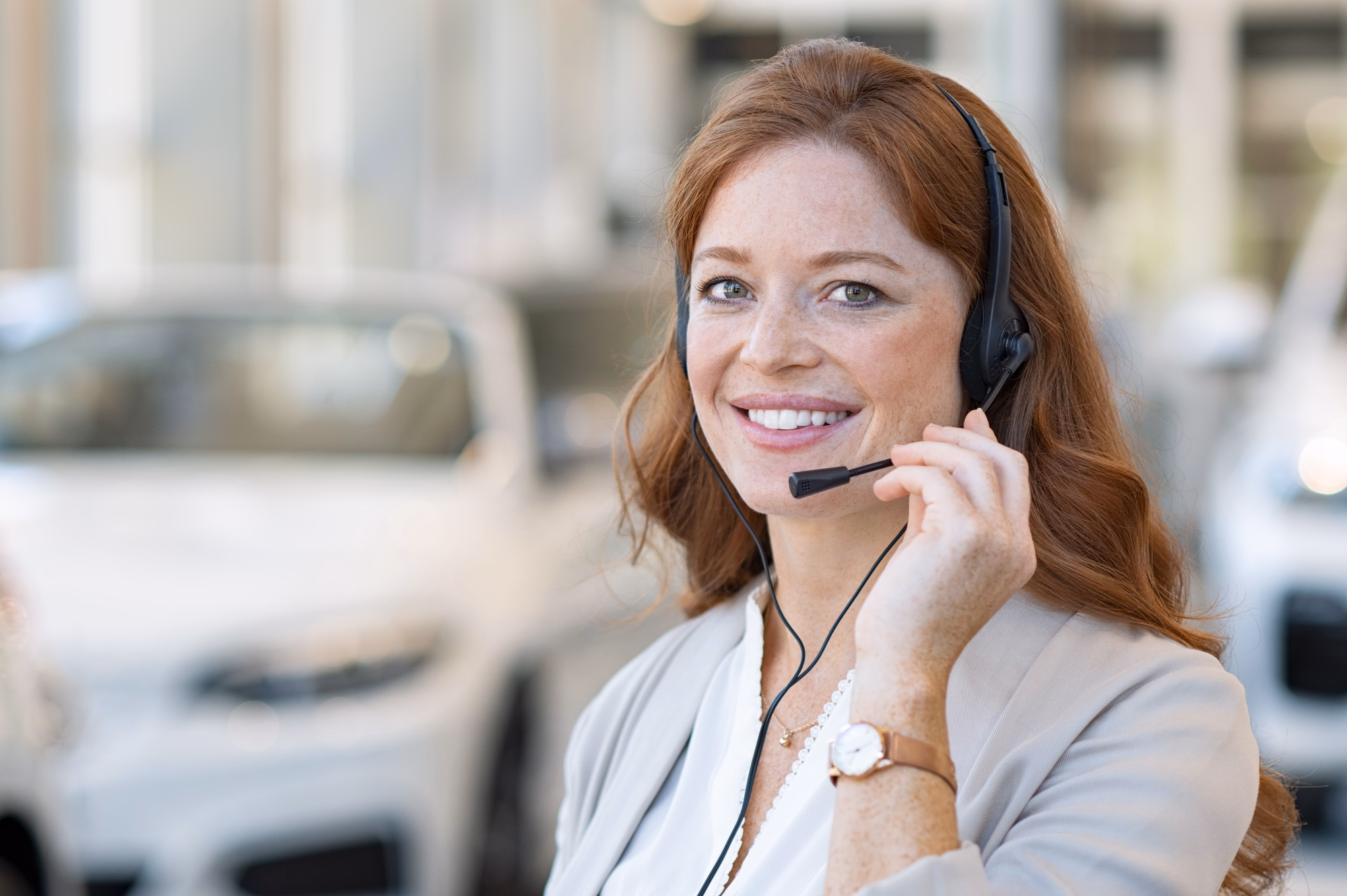 Beautiful customer support wearing headset and looking at camera. Portrait of smiling woman customer service representative working with cars in background. Call enter operator at car rental office.