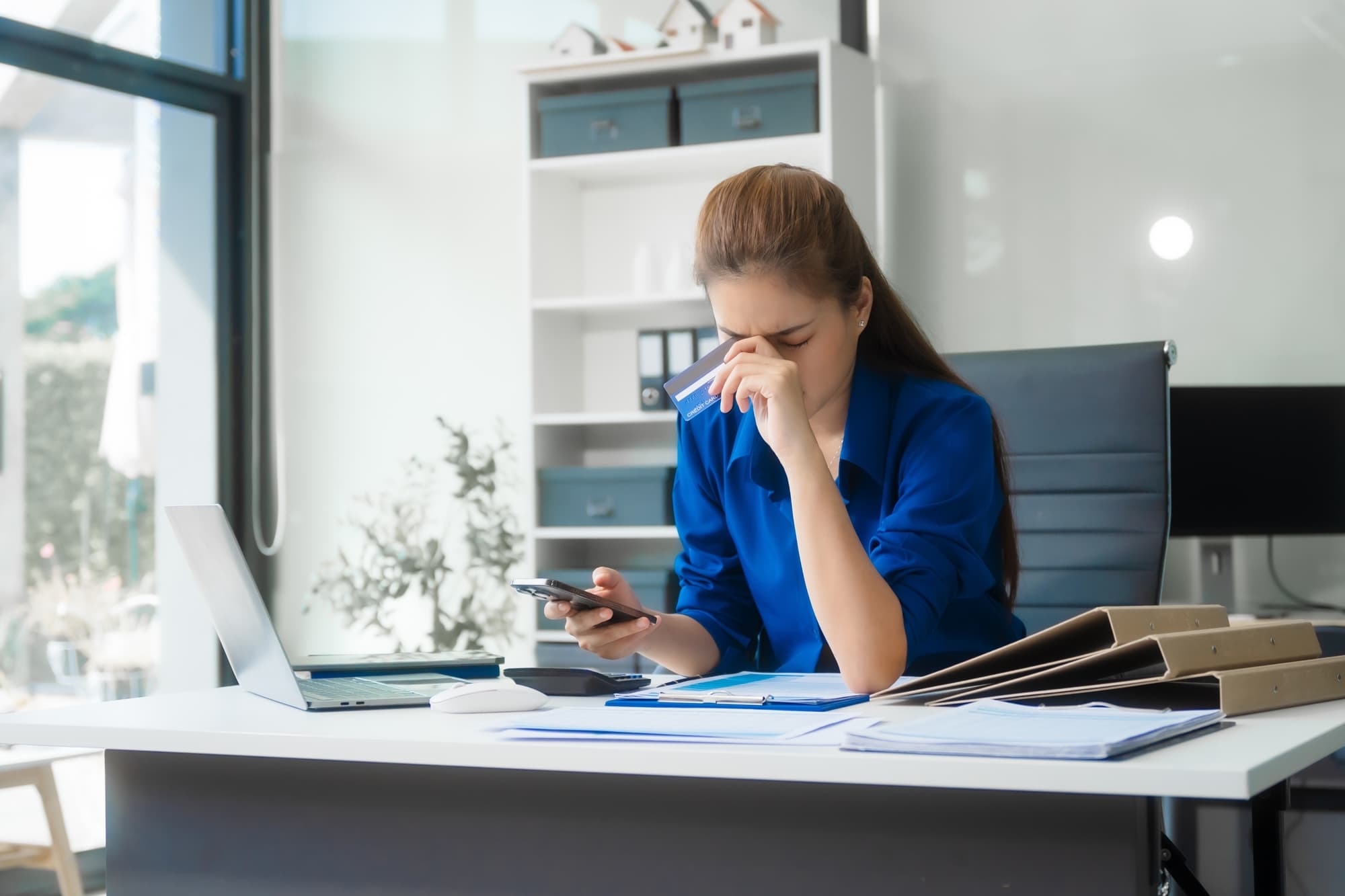 An Asian businesswoman in a blue formal shirt is working at her desk, analyzing financial charts on a laptop and mobile phone, focusing on credit management and financial strategies.