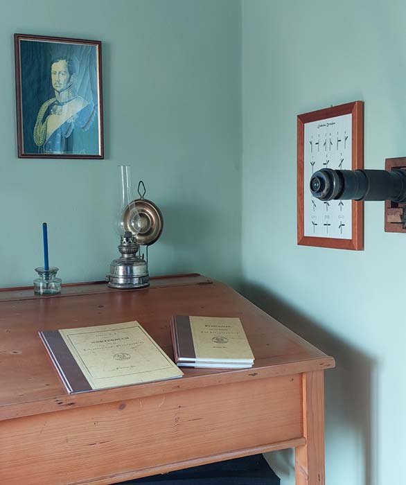 Holzschreibtisch mit zwei alten Büchern, einer Petroleumlampe, einem blauen Kerzenhalter, einem Portraitgemälde an der Wand und einem antiken Fernrohr neben einer Tafel mit Symbolen.