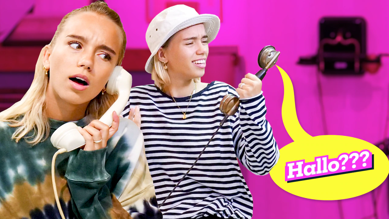 Lisa und Lena mit Telefonen, eine schaut verwirrt und hält einen weißen Hörer, die andere trägt ein gestreiftes Shirt und Hut, hält ein altes Telefon mit Sprechblase 'Hallo???', vor pinkem Hintergrund.