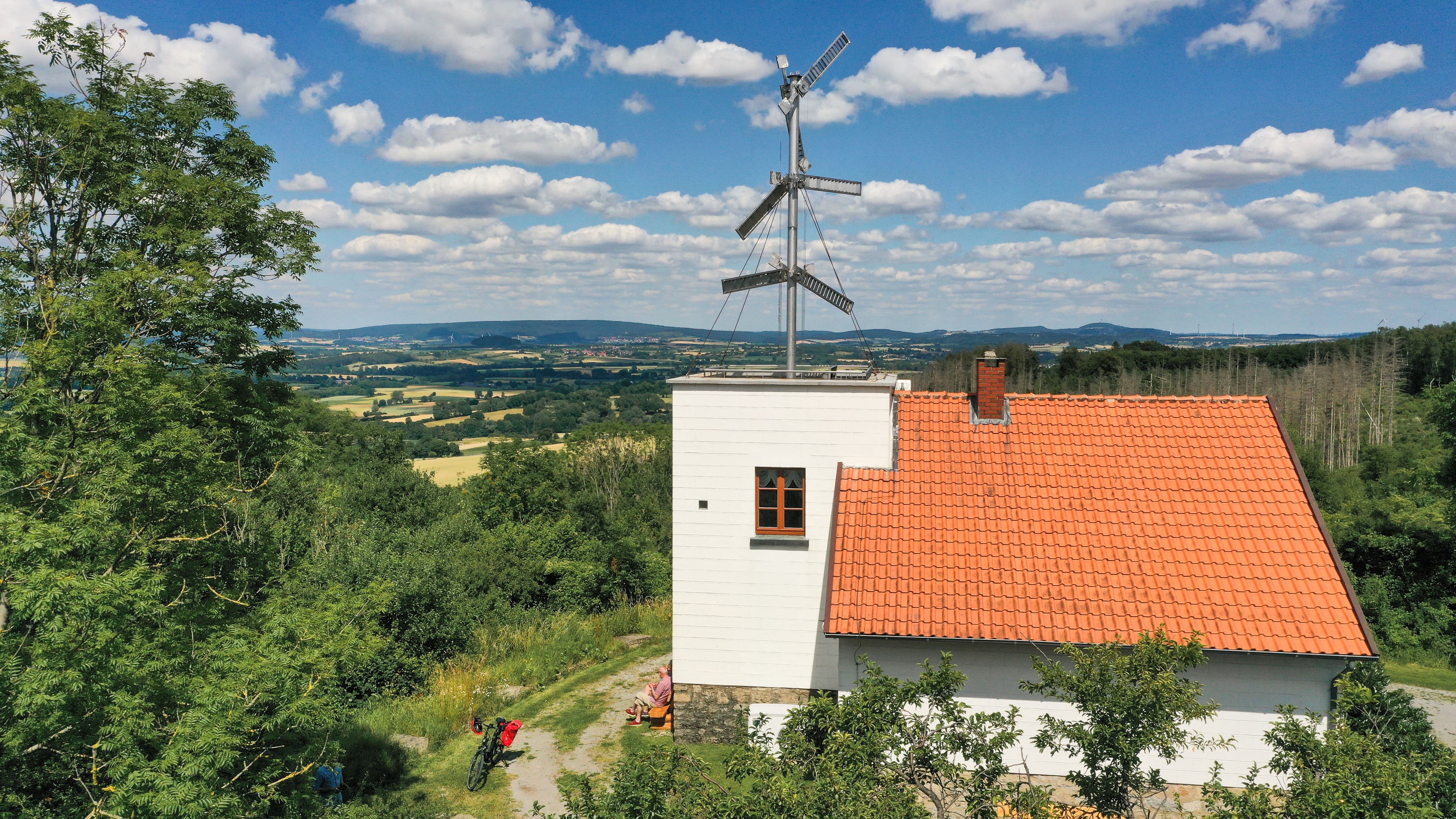 Telegrafenstation 32 mit einem Fahrrad davor, umgeben von grünen Bäumen und einer weiten Landschaft unter blauem Himmel mit Wolken.