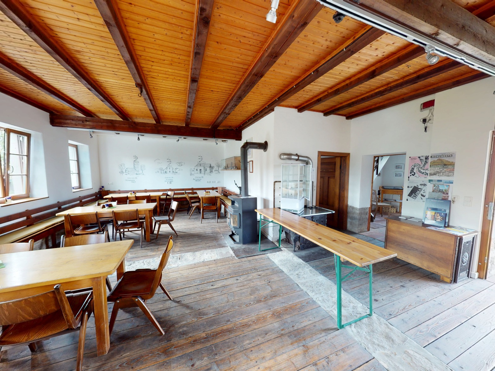 Gemütlicher Gastraum mit Holzboden, Holzdeckenbalken, mehreren Holztischen und -stühlen sowie einem Holzofen.
