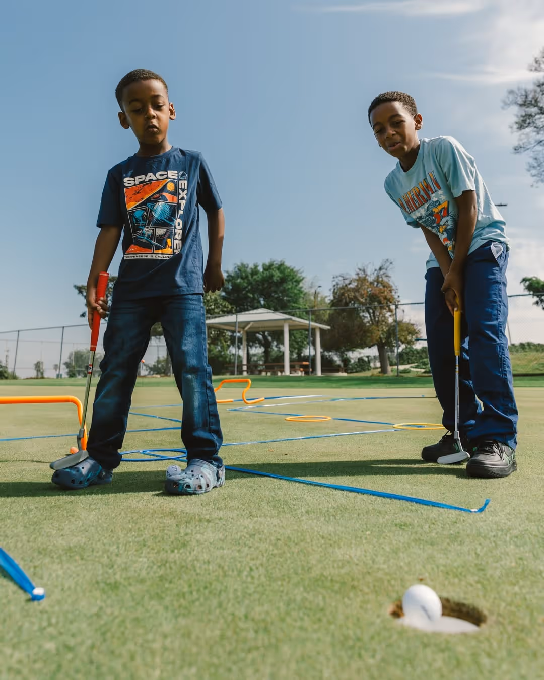 Two boys playing mini golf outdoors on a sunny day with golf balls near the hole.
