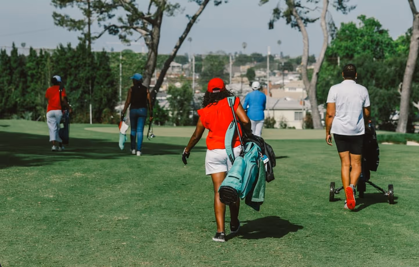 Five golfers walking on a green golf course carrying bags and pushing carts on a sunny day.