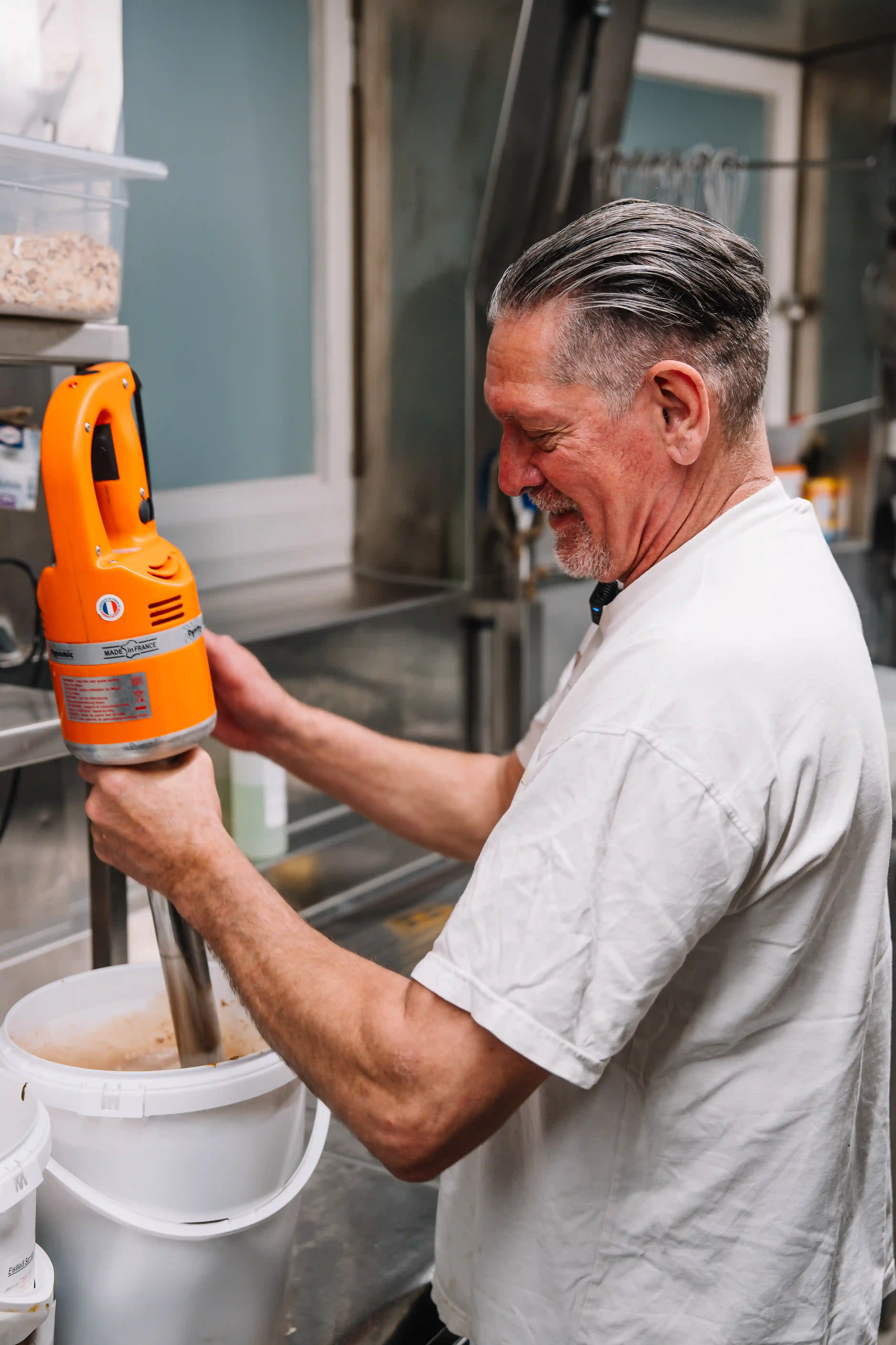 Man using an orange immersion blender to mix contents in a white bucket in a kitchen.