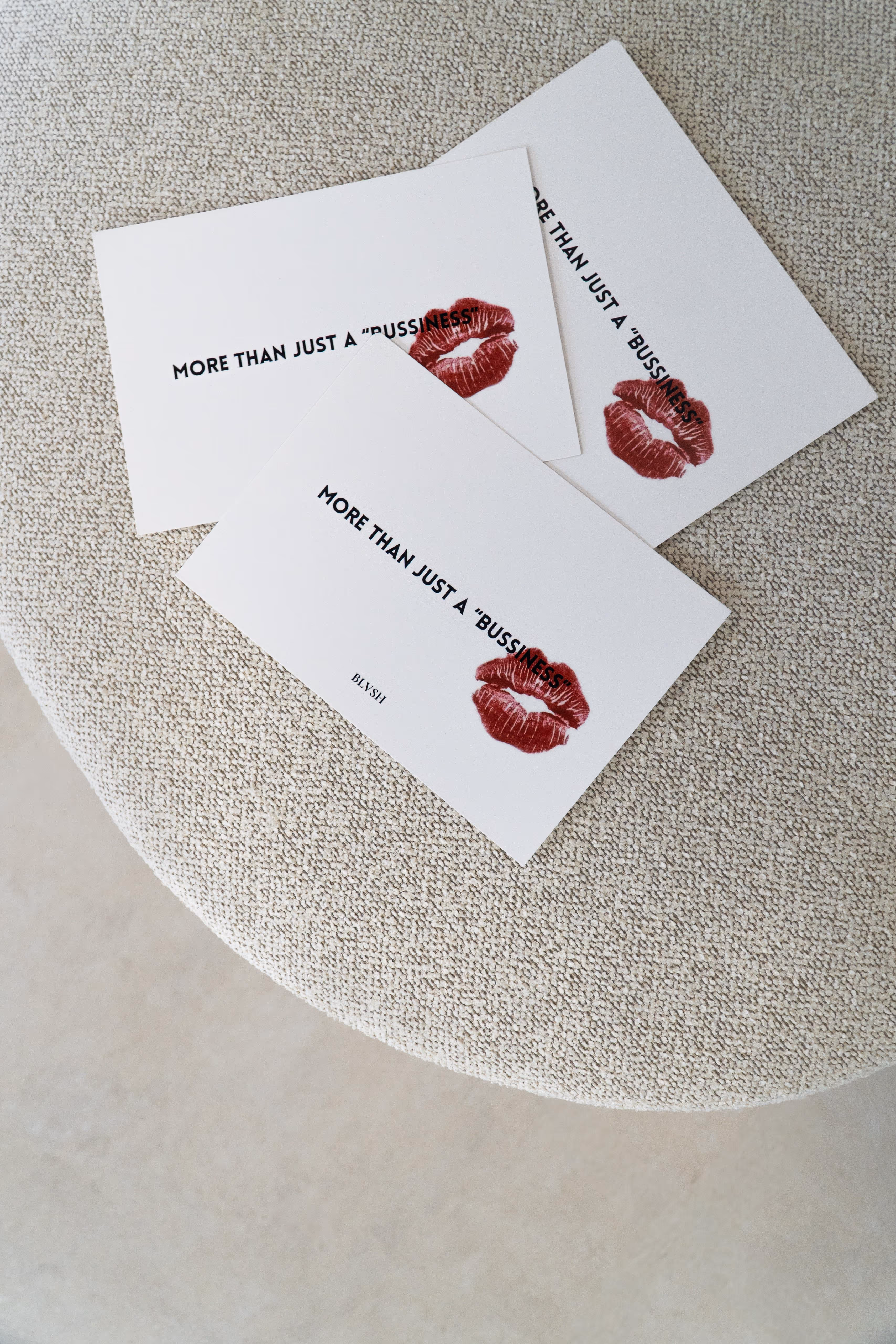 Three white cards with text ‘MORE THAN JUST A “BUSSINESS”’ and a red lipstick kiss on a textured fabric surface.