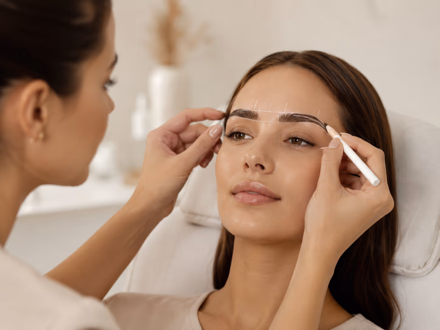 Woman getting eyebrow shape measured with white marker lines drawn on her face by a beautician.