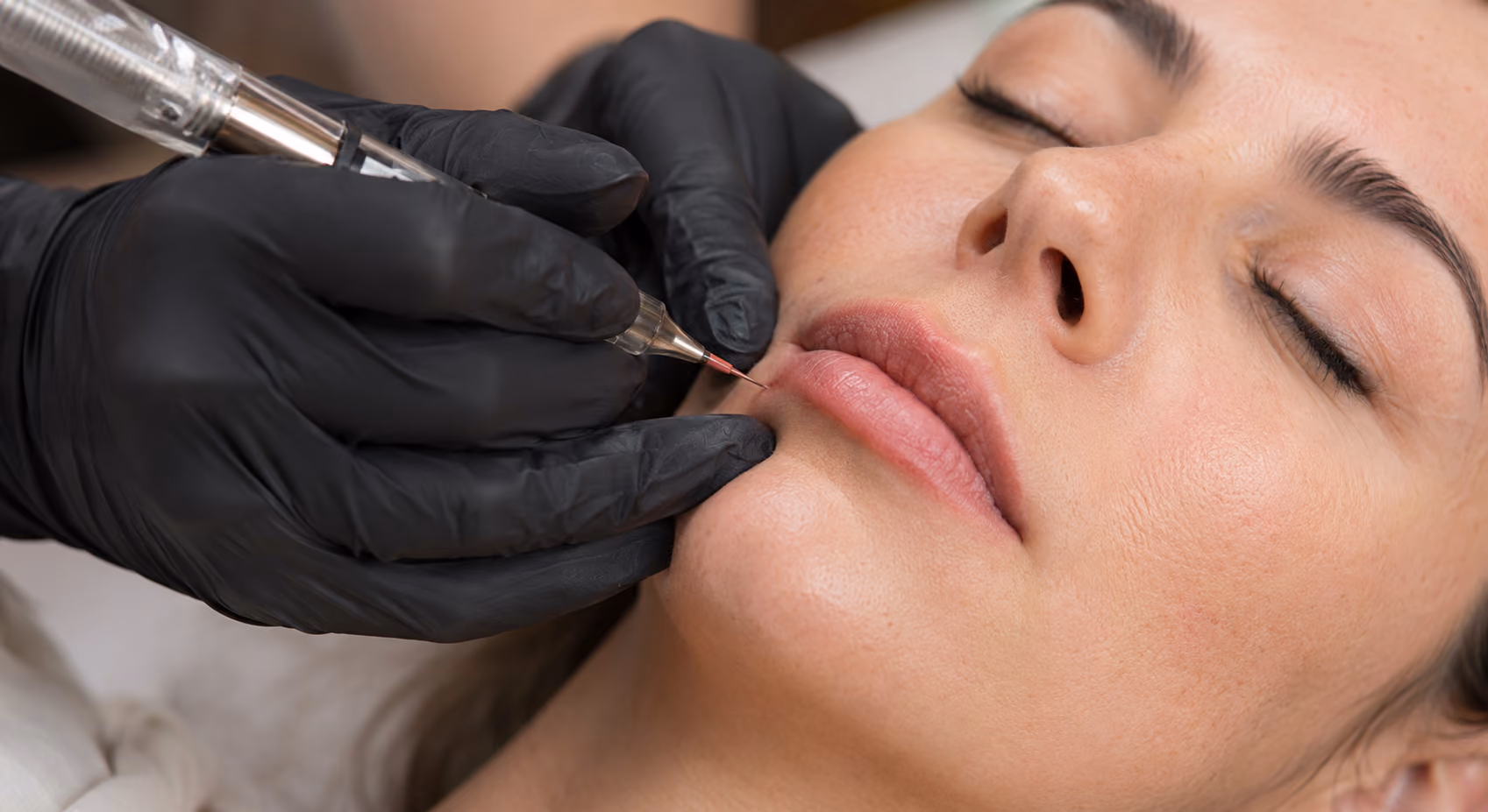 Close-up of a person receiving cosmetic microblading on their lips from a technician wearing black gloves.