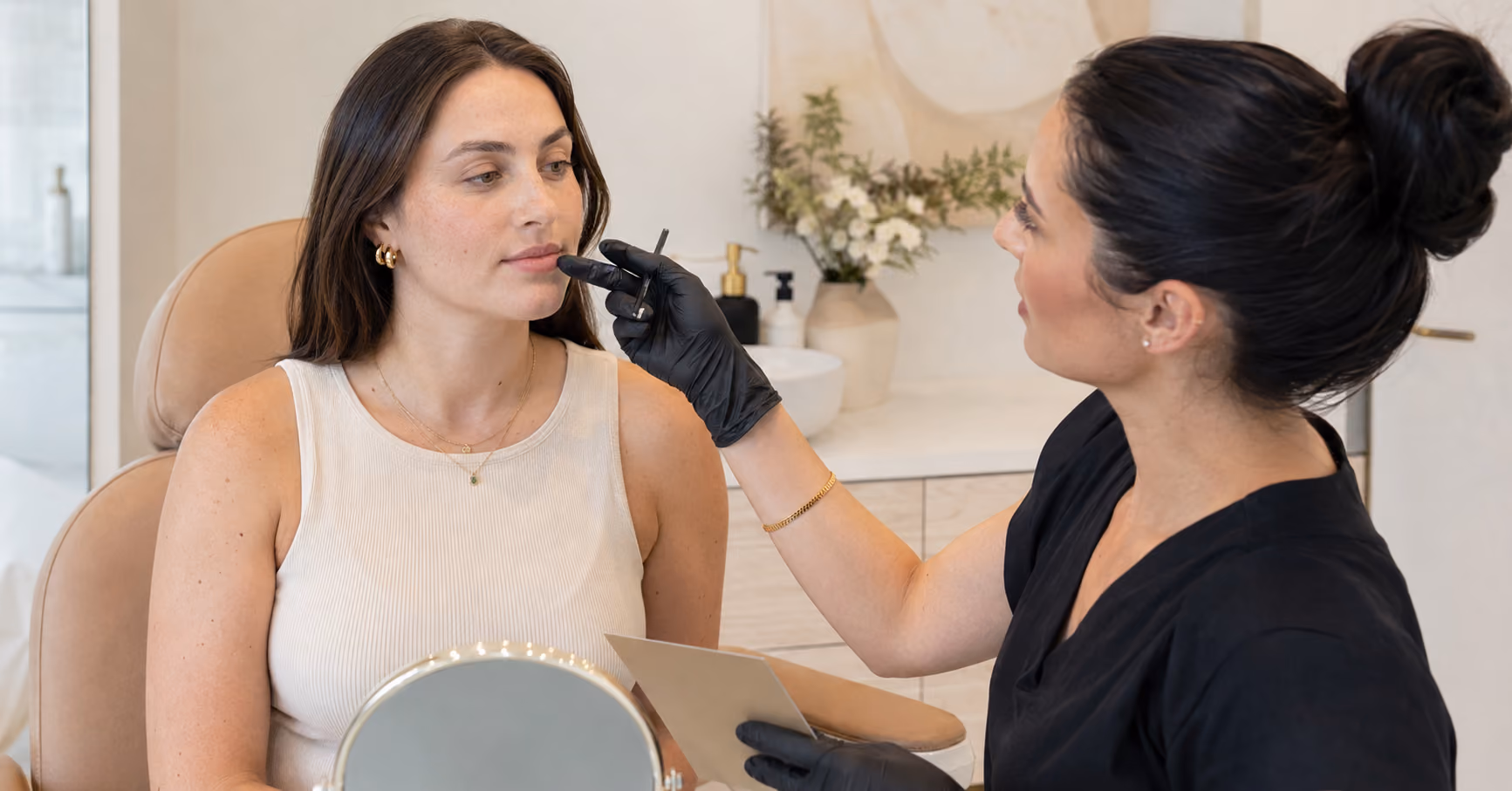 A beautician wearing black gloves applies lip makeup to a seated woman holding a mirror.