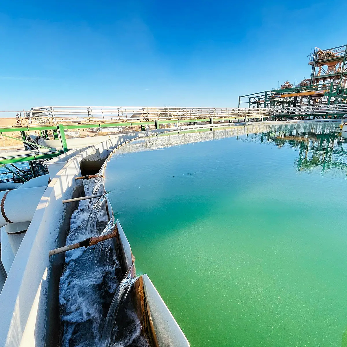 Circular industrial thickener tank with greenish water and water flowing over spillways under clear blue sky.