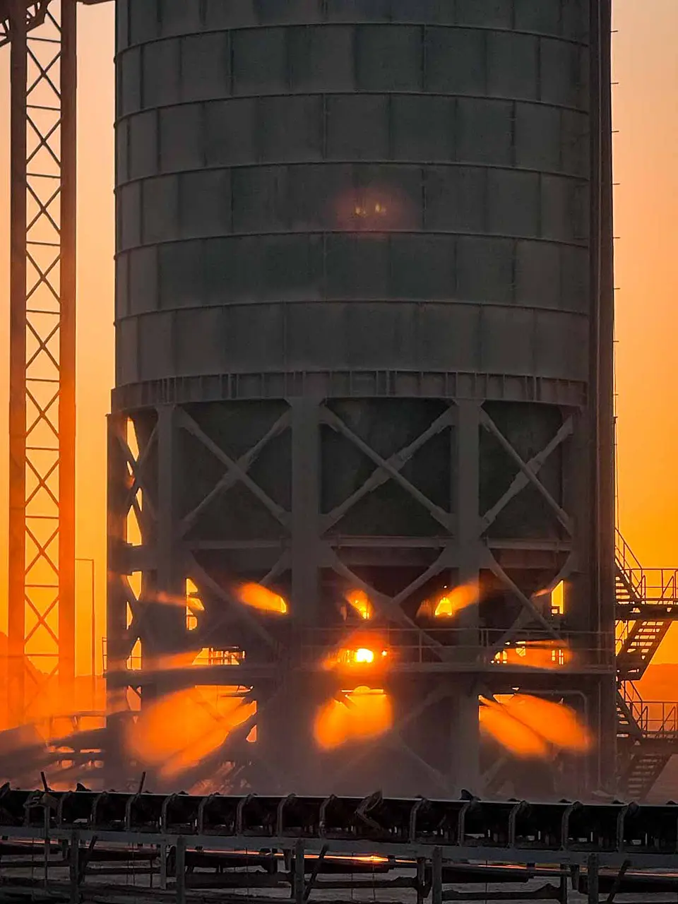 Industrial ore storage surge bin illuminated by orange sunlight shining through the lower structure at sunset.